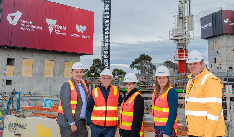 Five individuals in high vis orange vest and hard hats, in front of two cranes and a large concrete structure at the site of the new Footscray Hospital